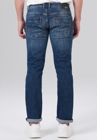 Jeans en denim bleu avec une coupe droite, comportant deux poches arrière, des coutures oranges et des ourlets roulés. Portés avec des baskets blanches.