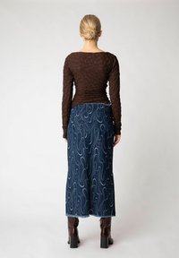 Brun tekstureret langærmet skjorte med samlede detaljer, parret med en lang denimnederdel med bølgede hvide mønstre og en frynset kant. Brun hæl-støvler.