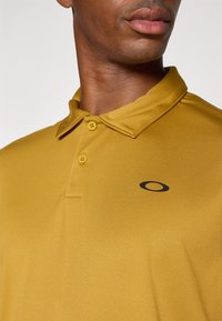Sinisen keltainen polo-paita, jossa on kaulus, kaksi nappia ja musta logo. Sileä rakenne, jossa on hieman kiiltoa.