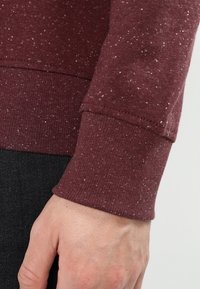 Detailní záběr na ruku vedle tkaniny v barvě hnědé s tečkami, se žebrovaným manžetovým lemem, nošené přes černé kalhoty.