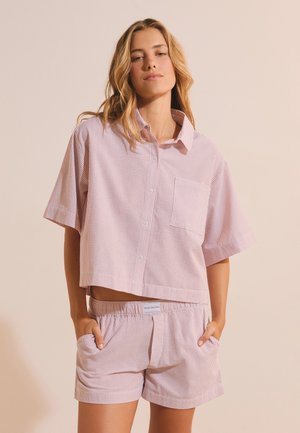 Vrouw draagt een losse roze-wit gestreepte korte mouwen blouse met voorzak en bijpassende short met elastische taille, handen in de zakken.