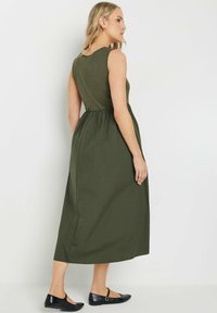 Vestido sin mangas en verde oliva con un corsé ajustado y una falda fruncida, que cae hasta la mitad de la pantorrilla. Combinado con zapatos planos negros.