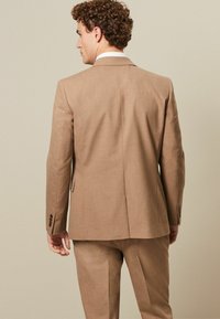 Brauner maßgeschneiderter Anzug mit Reverskragen und einer Schlitze hinten. Der Stoff wirkt strukturiert und weist geknöpfte Ärmelabschlüsse auf.