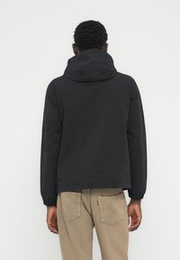 Μαύρο μπουφάν με κουκούλα και σχεδιασμό πλέγματος, ελαστικά μανίκια, και άνετη εφαρμογή, φορεμένο πάνω από μπεζ παντελόνι, με ματ υφή.