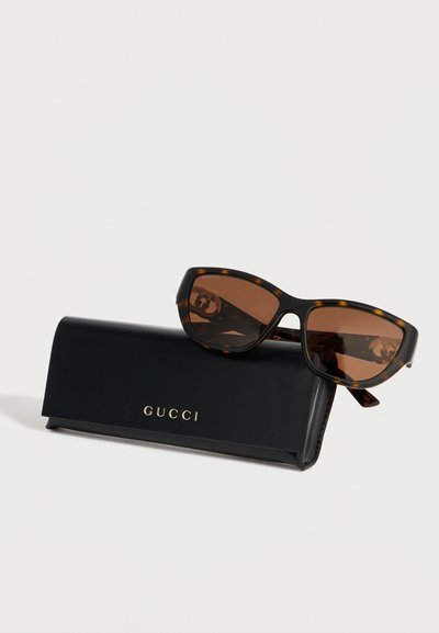 Γυαλιά ηλίου Gucci με καφέ φακούς και σκελετό χελώνας, που ακουμπούν σε μαύρη θήκη Gucci με χρυσά γράμματα σε λευκό φόντο.