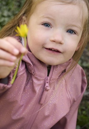 Bambino con giacca impermeabile rosa con pagliuzze dorate, che tiene in mano un fiore giallo. La giacca presenta un colletto alto e una chiusura con zip.