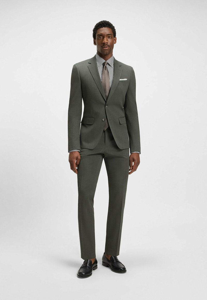 Dark green tailored suit with a textured finish, paired with a striped shirt and brown tie. Black loafers complete the outfit.