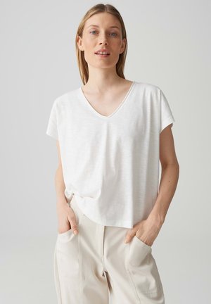 Frau mit heller Haut und blondem Haar trägt ein locker sitzendes weißes V-Ausschnitt-T-Shirt und hellbeige Hose, die Hände in den Taschen, vor schlichtem Hintergrund.