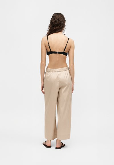 Γυναίκα με μακριά σκουρόχρωμα μαλλιά, ντυμένη με μαύρο σουτιέν, μπεζ φαρδιά παντελόνια και μαύρα σανδάλια slide, στέκεται γυρισμένη με την πλάτη προς ένα απλό λευκό φόντο.