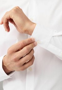 Chemise blanche en tissu lisse, avec des poignets boutonnés et un col arrondi. Une main ajuste le poignet, mettant en valeur un teint de peau naturel.