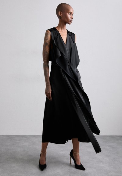 Black sleeveless dress with a draped front, made of smooth fabric. Features a slim waist and flowing skirt. Paired with pointed black heels.