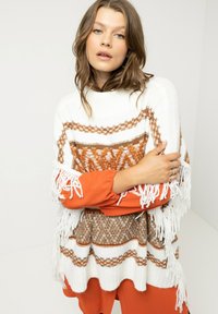 Weißes gestricktes Poncho mit braunen geometrischen Mustern und Fransen-Details; getragen über einem orangefarbenen Langarmoberteil, das einen kontrastierenden, geschichteten Textur-Effekt erzeugt.