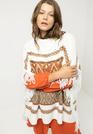 Witte gebreide poncho met bruine geometrische patronen en franjes; gedragen over een oranje langemouwen top, wat een gelaagd textuurcontrast creëert.