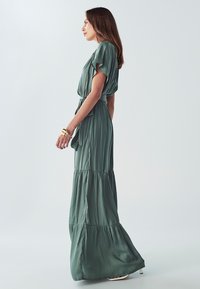 Dlouhé, šalvějově zelené šaty s krátkými rozšířenými rukávy, s vázačkou v pase a skládánou sukní. Vyrobeny z měkké, splývavé látky, mají hladký povrch.