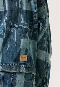 Džínová bunda s abstraktními modrými a černými vzory nastříkanými sprejem a malou hnědou štítkem "Pepe Jeans London" na zadním lemu.