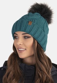 Bonnet en tricot teal avec un motif en relief de câble, ornée d'un grand pompon en fausse fourrure noire sur le dessus et d'un petit accent en cuir avec le logo.