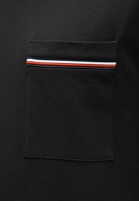 Μαύρο ύφασμα με υφή που διαθέτει τσέπη με πατς, στολισμένη με φερμουάρ σε ρίγες κόκκινου, λευκού και μπλε κατά μήκος της κορυφαίας άκρης.