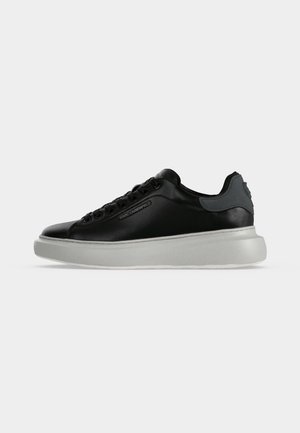 Zwarte lage sneaker met zwarte veters en grijze hielpatch, met dikke witte zool en subtiele branding aan de zijkant.