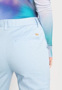 Vaaleansiniset housut, joissa on sileä pinta, takatasku ja pieni kultaiseen sävyyn sopiva koriste.