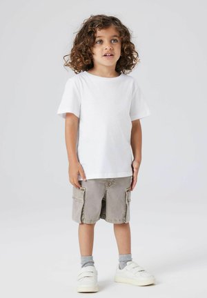 Ung barn med krøllete hår iført hvit t-skjorte, beige cargoshorts, grå sokker og hvite joggesko som står mot en ensfarget bakgrunn.