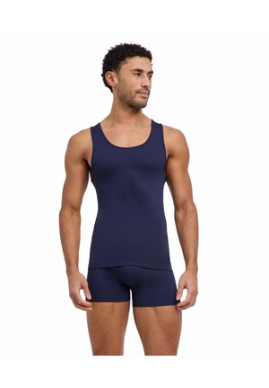 Atletische man in marineblauw tanktop en korte broek, staat met ontspannen handen en kijkt iets naar de zijkant tegen een witte achtergrond.