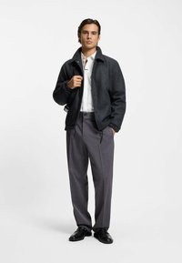 Dunkelgraue Wolljacke mit klassischem Kragen, kombiniert mit texturierten grauen Hosen mit einem dezenten Karomuster. Schwarze Schuhe runden den Look ab.