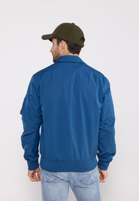 Veste bombers bleue en tissu lisse avec des poignets et un ourlet côtelés. Associe avec un jean bleu clair et une casquette vert olive.