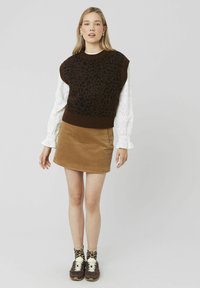 Un gilet sans manches à imprimé léopard marron porté sur une chemise à manches longues en dentelle blanche, associé à une mini-jupe en velours côtelé beige et des baskets marron.
