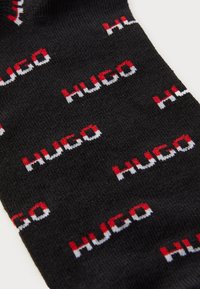 Schwarze Socken mit gewebtem "HUGO"-Text in rot, weiß und schwarz, mit einem sich wiederholenden Muster über den Stoff. Weiche Textur.