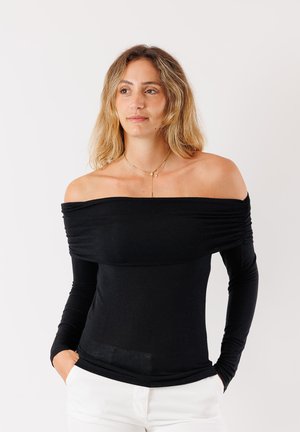 Top nero scollato con maniche lunghe, caratterizzato da un tessuto arricciato sulla zona del torace e una texture liscia. Abbinato a pantaloni bianchi.
