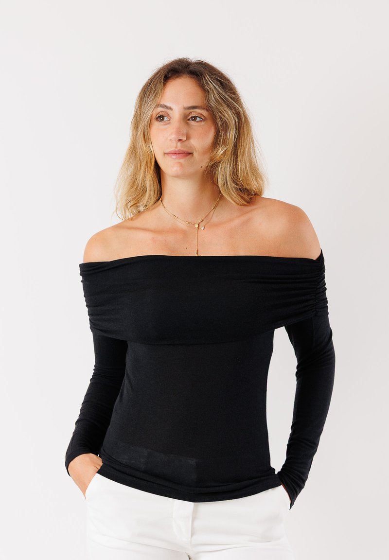 Top nero scollato con maniche lunghe, caratterizzato da un tessuto arricciato sulla zona del torace e una texture liscia. Abbinato a pantaloni bianchi.