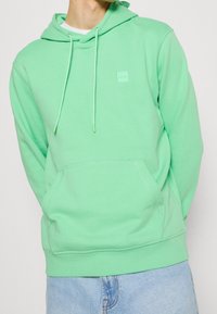 Felpa verde in morbido tessuto, con tasca frontale, cappuccio regolabile e una piccola etichetta del brand sul petto. Vestibilità casual.