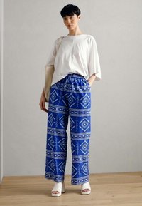 Un haut blanc surdimensionné associé à un pantalon large bleu à motifs doté de designs géométriques et d'une taille élastique, complété par des sandales à talons blocs blanches.