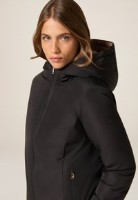 Czarna ocieplana kurtka z kapturem, z zapięciem na zamek błyskawiczny z przodu, bocznymi kieszeniami i gładką fakturą. Minimalistyczny design bez widocznych wzorów czy logo.