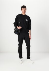 Sweatshirt preto de gola redonda com um gráfico, calças pretas ajustadas e ténis brancos. O modelo leva uma pequena bolsa preta com detalhes metálicos.
