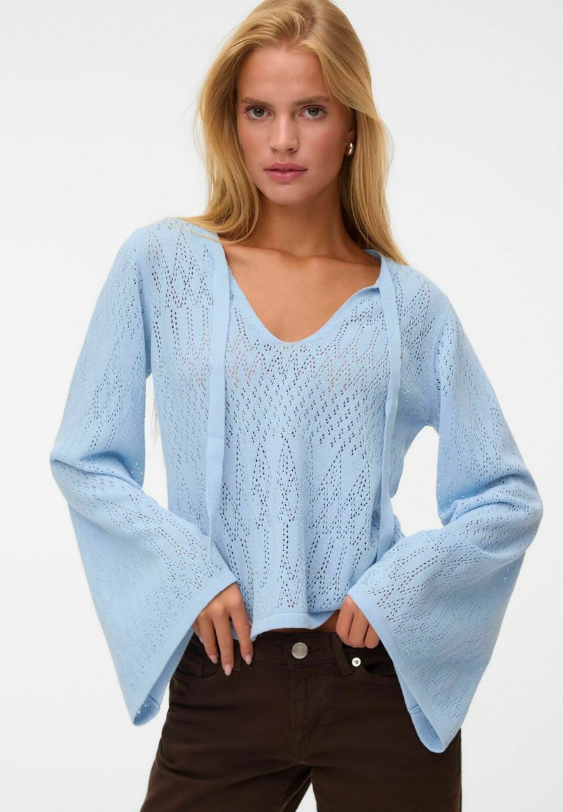 Femme blonde portant un pull en maille bleu clair à col en V avec de larges manches et un pantalon noir, debout devant un fond blanc uni.