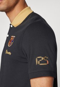 Homme portant un polo noir avec un col beige, affichant le blason du FC Barcelone, le logo Spotify et un emblème du 125e anniversaire sur la manche.