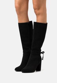 Botas altas de ante negro, con tacón ancho, que cuentan con una cremallera lateral y un detalle de lazo en la parte superior. Textura suave y diseño streamlined.
