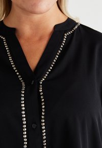 Blusa preta com decote em V, apresentando uma fileira de tachas decorativas em tom prateado na frente. Tecido suave com uma textura lisa.