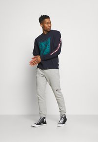 Maglione navy con grafica turchese e testo rosa sulle maniche, abbinato a pantaloni della tuta grigi chiari e scarpe nere alte. Tessuto di pura morbidezza.