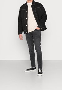 Giubbotto in denim nero con cuciture bianche, felpa beige, jeans grigio scuro e scarpe da ginnastica nere con suola bianca. Outfit semplice e casual.