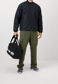 Giacca bomber nera, pantaloni cargo verde oliva, scarpe sportive grigie, borsa a tracolla nera con striscia bianca e logo, tenuta con una mano.
