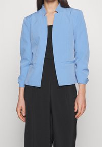 Světle modrý blazer s vysokým límcem, dlouhými rukávy a strukturovaným designem. Obsahuje dekorativní švy a dvě přední kapsy.