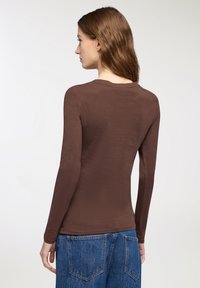 Donna con lunghi capelli castani, che indossa una camicia aderente marrone a maniche lunghe e jeans di denim blu, in piedi di spalle su uno sfondo bianco.