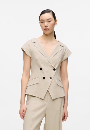 Kobieta ubrana w beżową, krótka marynarkę dwurzędową z czarnymi guzikami oraz pasujące beżowe spodnie, stojąca na białym tle.