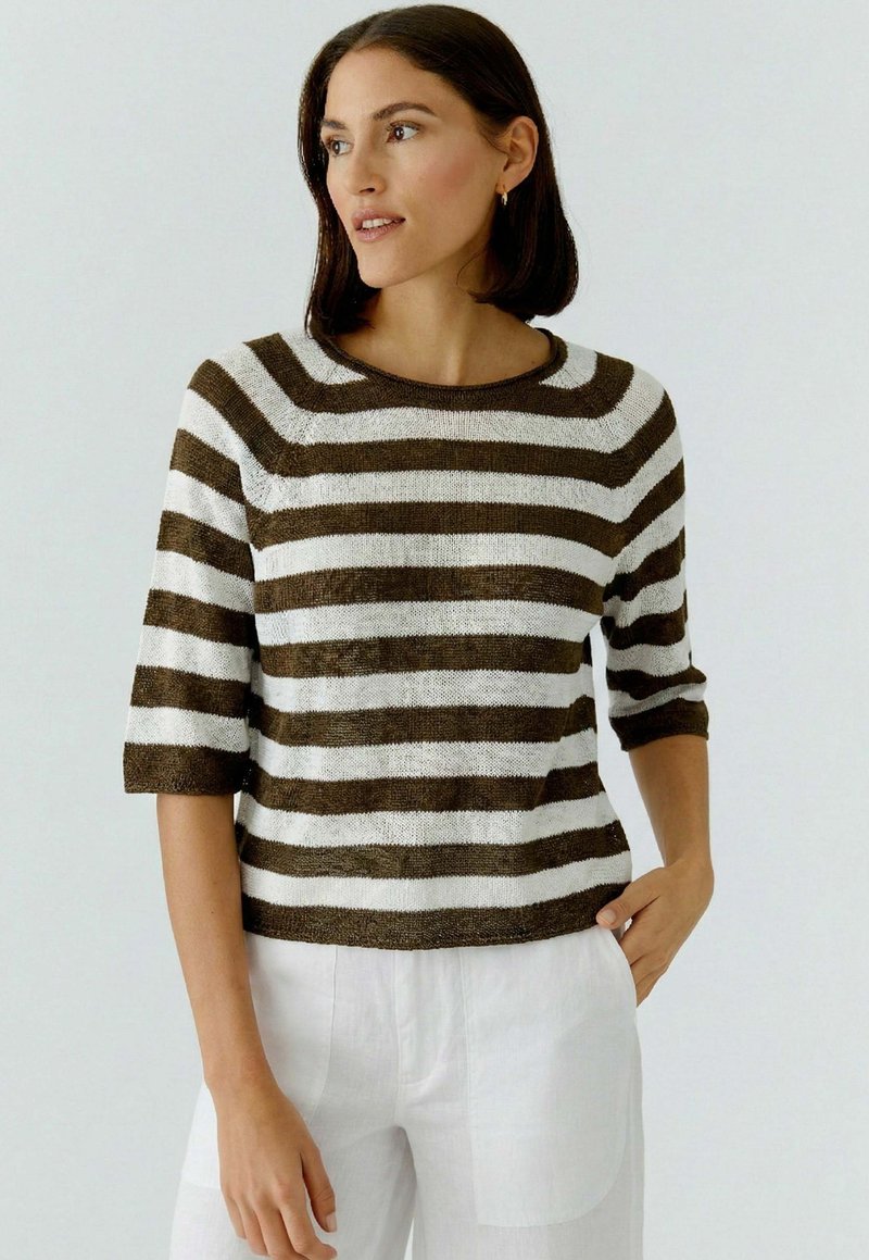 Femme aux cheveux foncés mi-longs portant un pull à manches courtes rayé marron et blanc et un pantalon blanc, debout avec une main dans la poche.