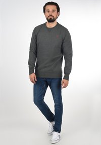 Grijze sweatshirt met ronde hals en rood logo, gecombineerd met blauwe jeans en witte sneakers. Heeft ribbelboorden en een gladde textuur.
