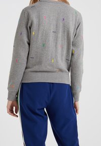 Persona que lleva un suéter gris con pequeños logotipos bordados de colores y la palabra "POLO", combinado con pantalones azules con franjas a los lados.