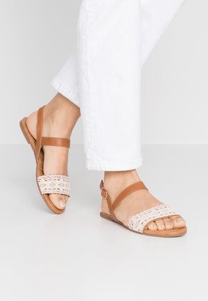 Brune lædersandaler med en snor mønstret beige rem. Har en justerbar ankelrem og flad sål. Båret med hvide bukser.