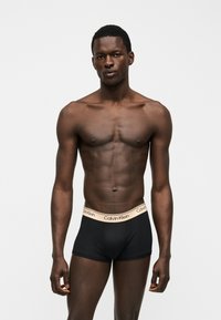 Mandlig model med mørk hud, iført sorte Calvin Klein boxershorts med beige linning, stående mod en ensfarvet hvid baggrund.